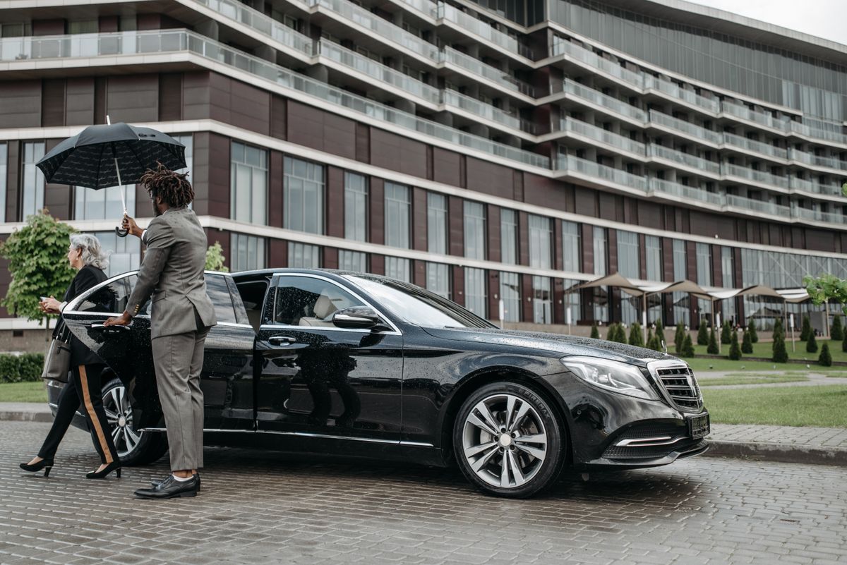 Chauffeur in pak houdt een paraplu boven een passagier bij een donkere personenauto